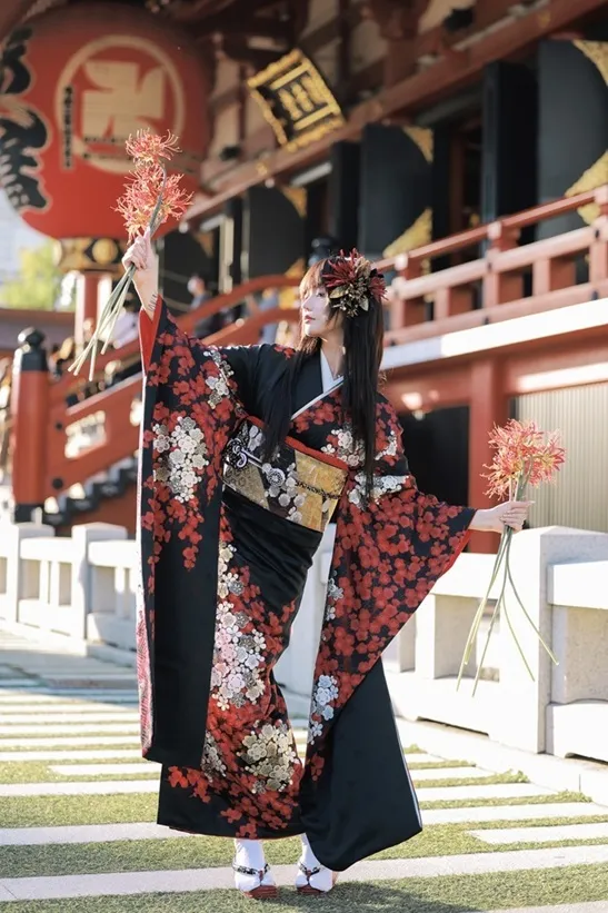 Full-length view at Sensoji of a woman in a black and red floral furisode holding spider lilies